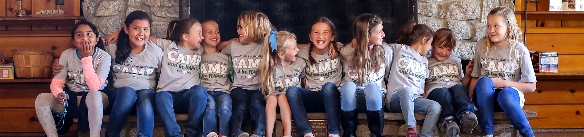  eleven girls sitting on a hearth in front of a fireplace in the lodge at camp skyland ranch with their arms around each other smiling and laughing 