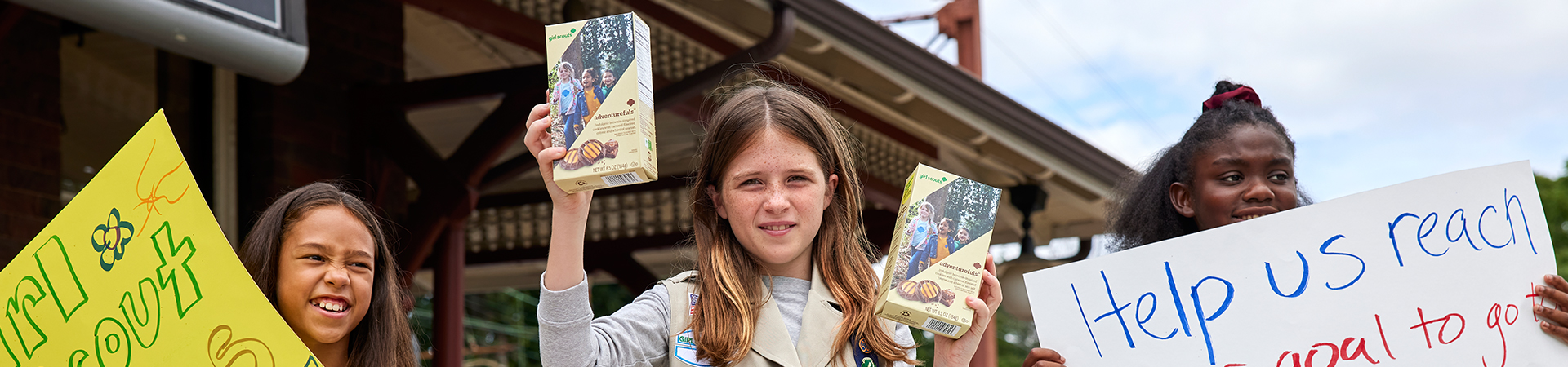  three girls holding signs and girl scout cookie boxes at a cookie booth 