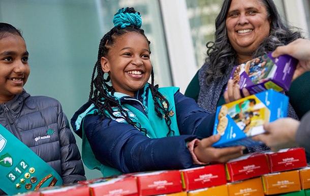 Two girls and an adult at a cookie booth with one girl handing two boxes of cookies to an unpictured adult.