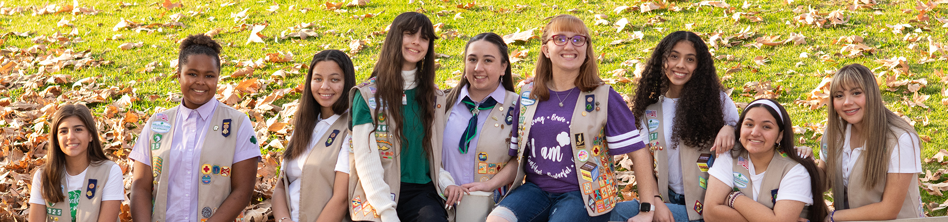  photograph of 9 older Girl Scouts smiling in front of a grassy hillside with fall leaves 